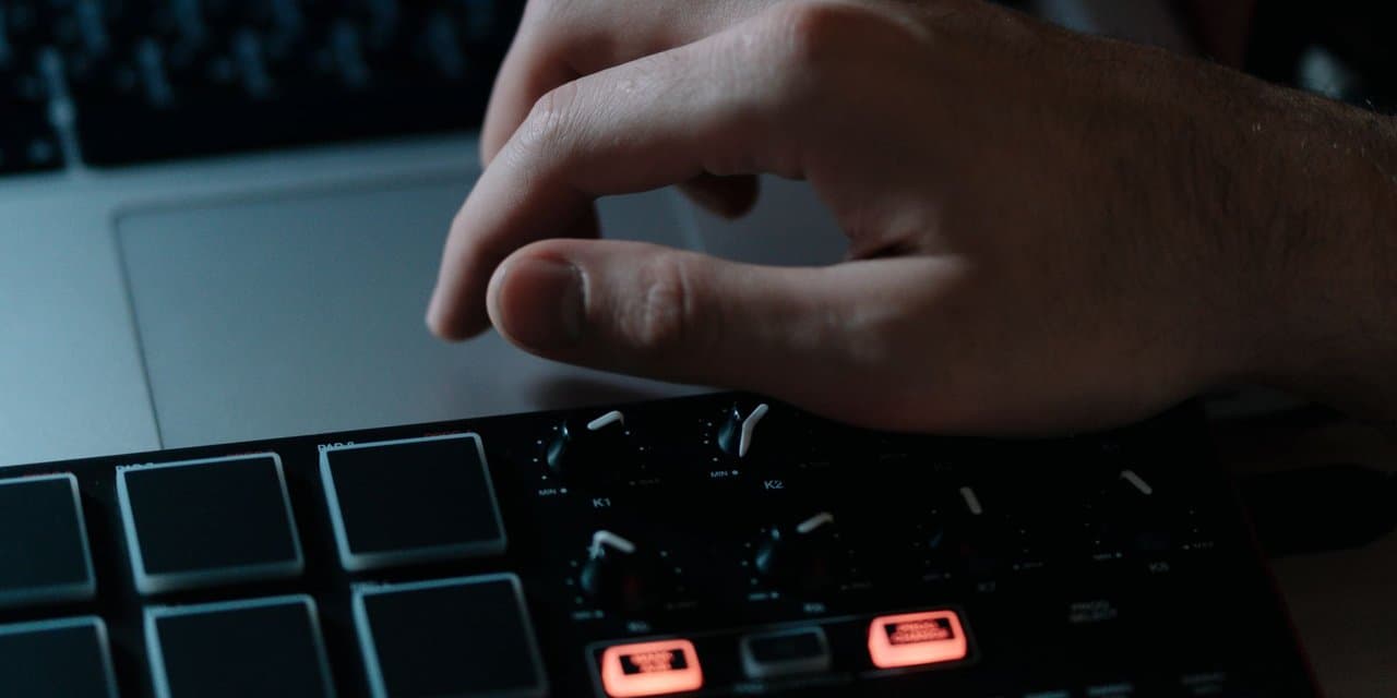 Tangan produser di atas pad drum dan knob MIDI controller di depan laptop, dalam pencahayaan studio yang gelap