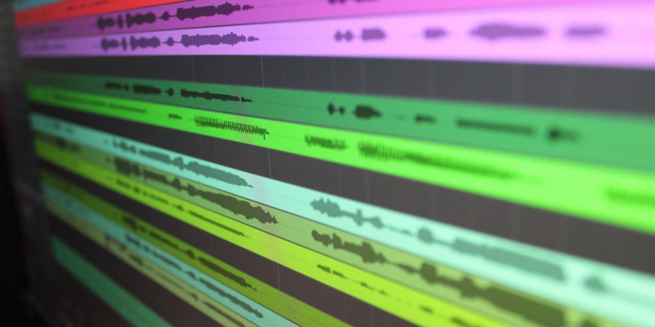 Close-up layar DAW menampilkan beberapa track audio berwarna-warni dengan waveform bertumpuk, menunjukkan proses layering instrumen saat aransemen musik