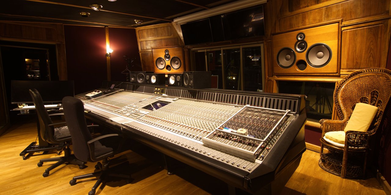 Ruang kontrol studio rekaman dengan mixing console analog besar, dua kursi operator, dan monitor speaker terpasang di panel kayu