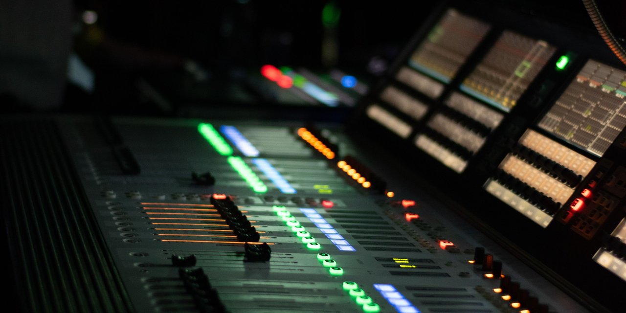Close-up mixing console dengan lampu indikator hijau, biru, dan oranye menyala di ruang studio gelap